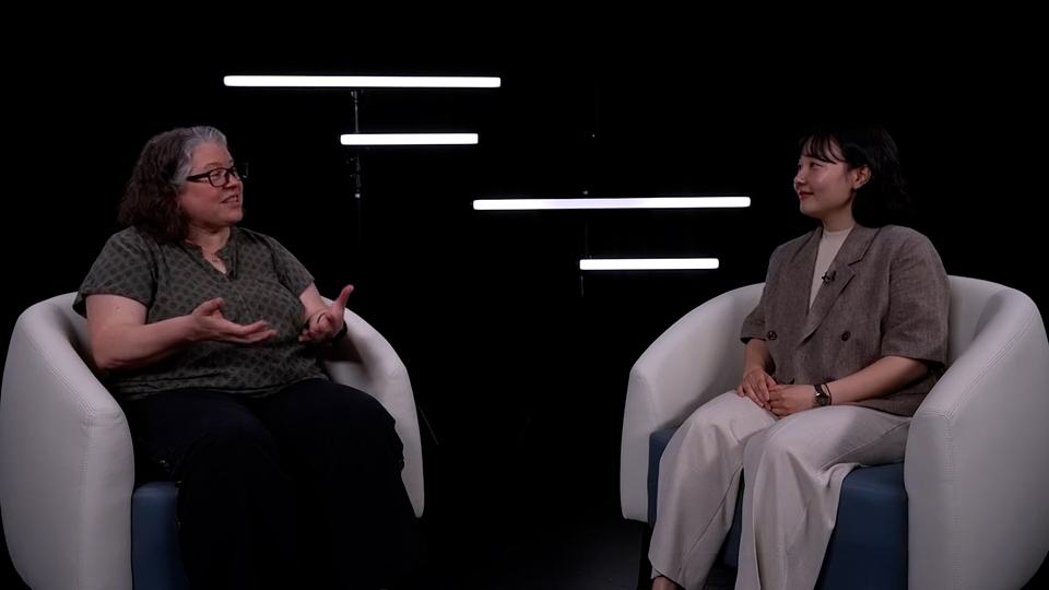 Two women sit facing each other in white armchairs during a studio interview against a black background with horizontal light bars.