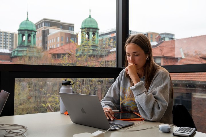 A student sits at a table near a large window, concentrating on a laptop while holding her hand near her chin. A tablet, water bottle, calculator, and wireless earbuds rest on the table, and historic buildings with green domed towers are visible outside.