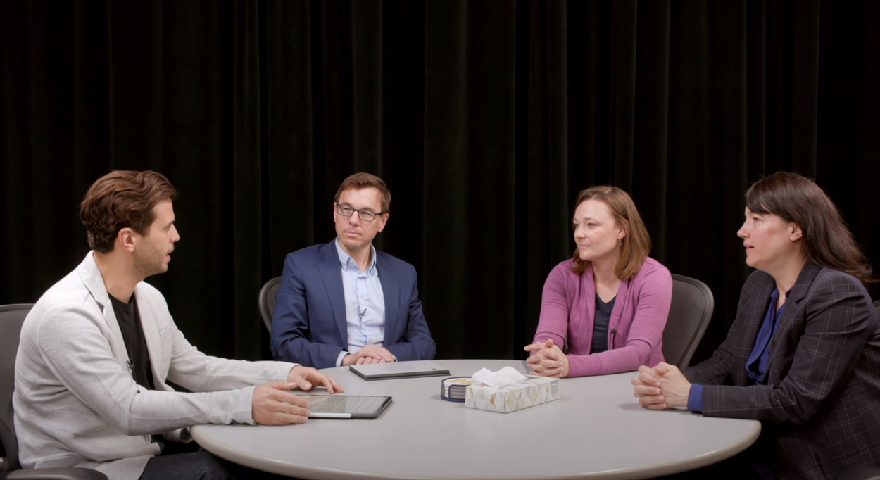 Four people sit around a circular table in discussion. One person is speaking; others listen attentively.