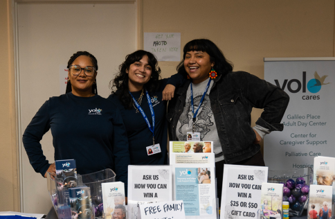 Aliya Patel smiles with coworkers stand together behind a table displaying brochures, sign-up sheets, and small giveaway items. A banner behind them reads “Yolo Cares” and lists services including caregiver support and hosp