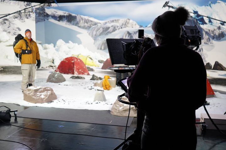 A man wearing a yellow jacket, winter hat, gloves, and a backpack stands in a staged snowy campsite with colorful tents and artificial snow. In the foreground, a camera operator in dark clothing films him, with studio lights and equipment visible.