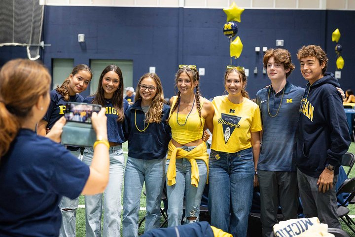A group of students in U-M gear smile and wave at the camera